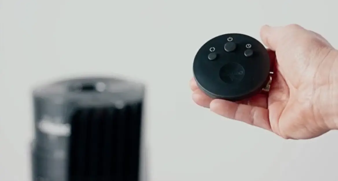 OSC54 Remote A person holding the OSC54's remote in their hand. The remote is circularly shaped with three gray buttons. The left button is for oscillation, the middle button is the power and fan speed button, and the right button is the timer. In the background is a blurred image of the OSC54 tower fan.