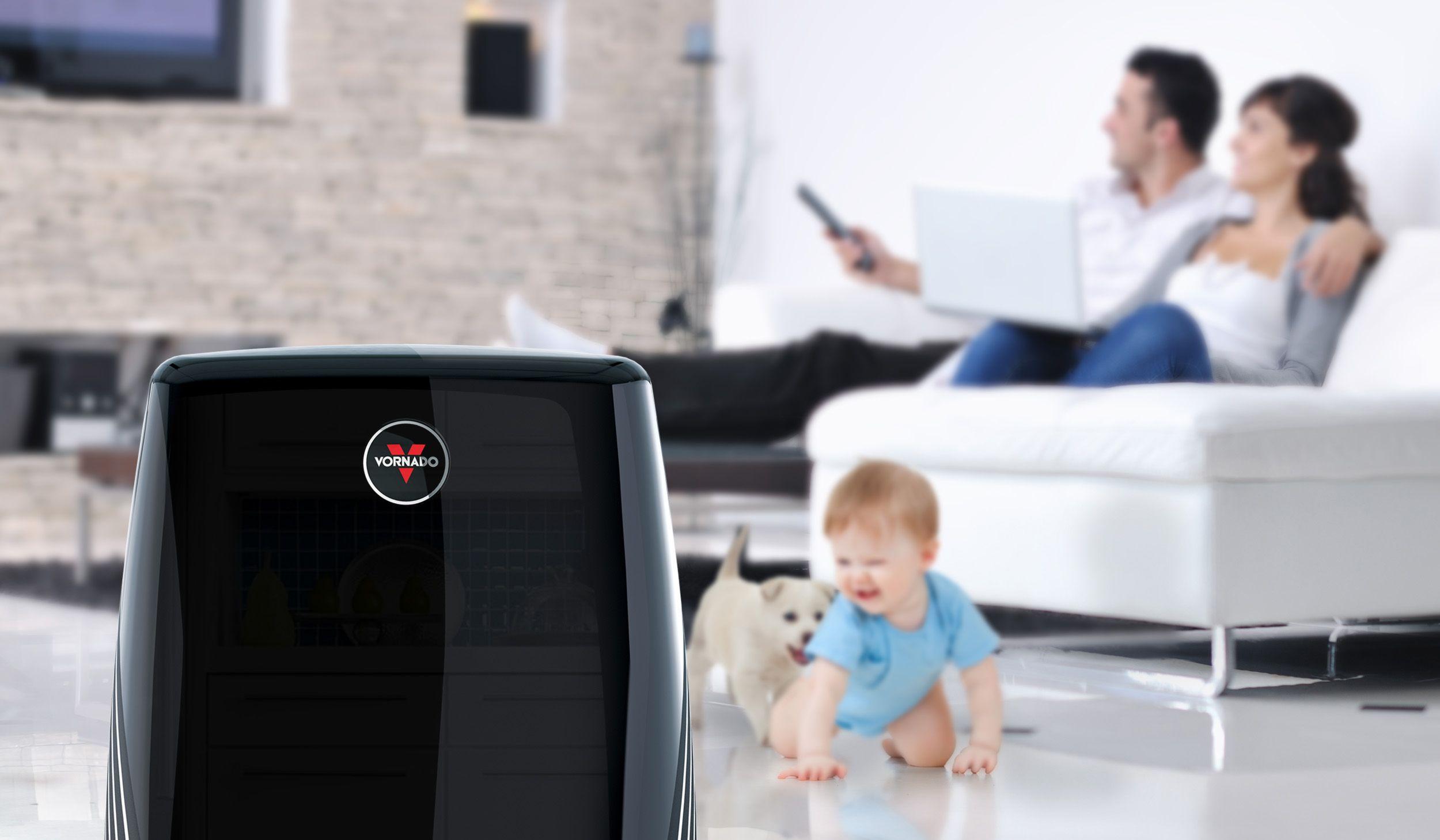 PCO300 Lifestyle Lifestyle of a black PCO300 air purifier in the foreground. In the background, blurred, is a crawling baby and a puppy. On the couch, the father is changing the channel on the tv and the mother is on her laptop.