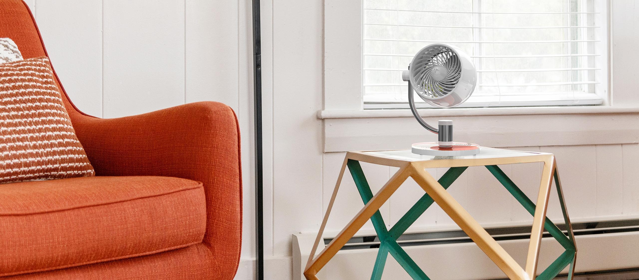 Pivot3U Lifestyle Banner Pivot3U Compact Air Circulator on a abstract coffee table with geometric legs. There is a bright red sofa next to the table.